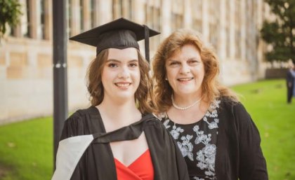 A young woman in graduation garb smiles next to an older woman in UQ's Great Court.